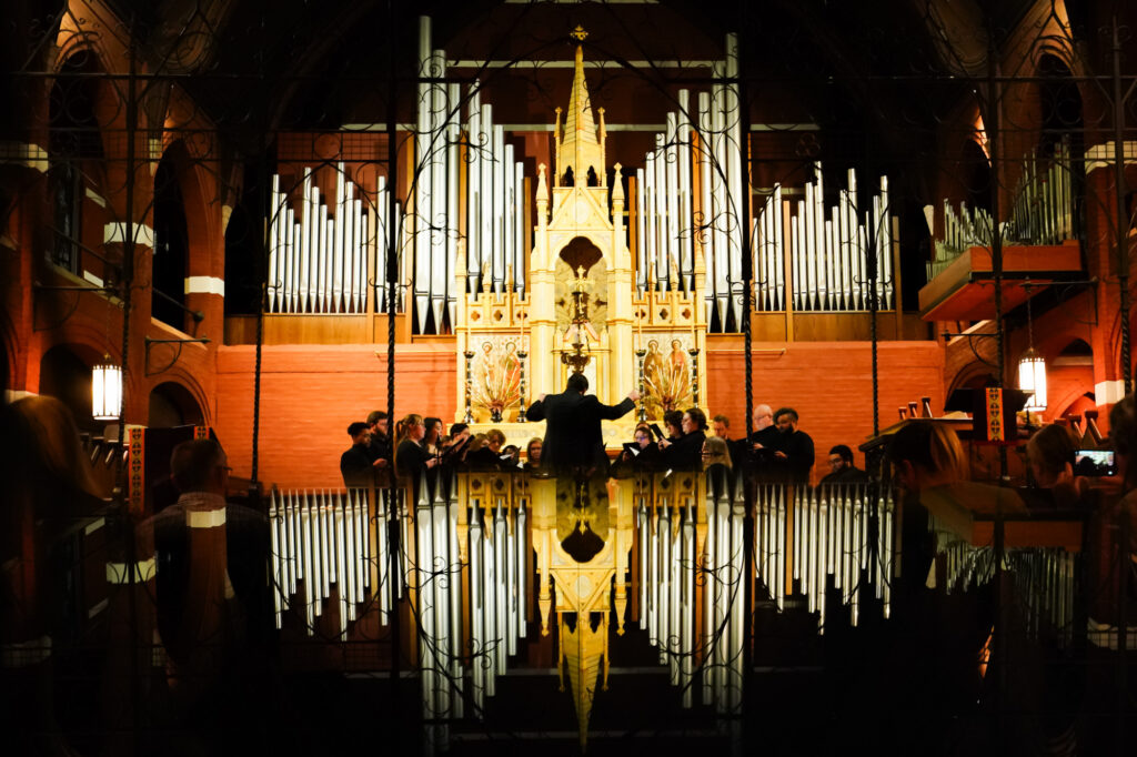 Choral performance reflected in a glossy dark surface as a choir sings in front of a grand pipe organ inside a brick chapel.
