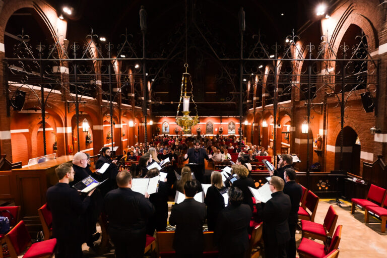 View from behind the choir as Dr. Anthony Maglione conducts a performance for an audience seated in a red brick chapel with wrought iron railings.