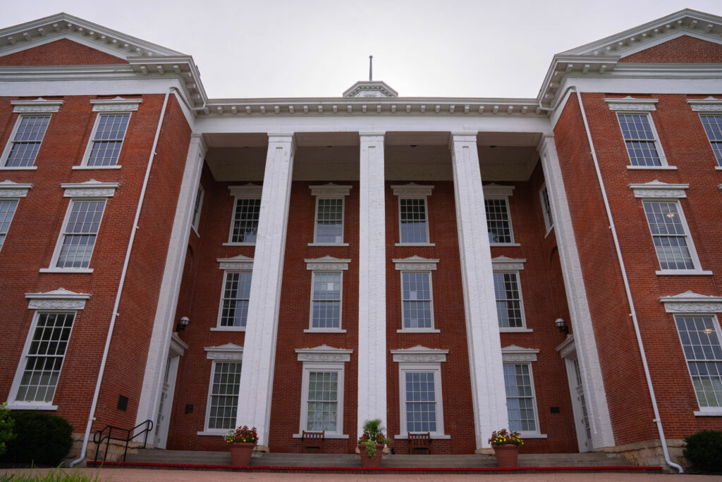 Low-angle exterior view of Brown Hall at William Jewell College, showcasing red brick walls and 4 white pillars.