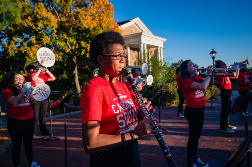Members of The Cardinal Sound marching band playing brass and woodwind instruments outdoors near Pillsbury Music Center.