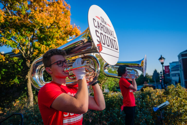 A student in a red shirt playing a marching tuba featuring a "The Cardinal Sound" logo on the bell cover.