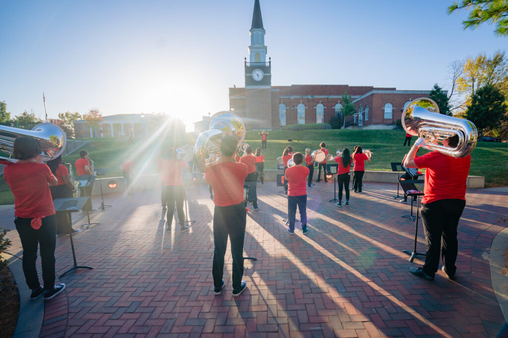 The Cardinal Sound marching band practicing on a brick plaza while facing Gano Chapel under bright evening sunlight.