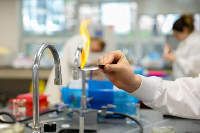 Close-up of a student’s gloved hand holding a metal strip over a flaming Bunsen burner during a chemistry lab experiment.