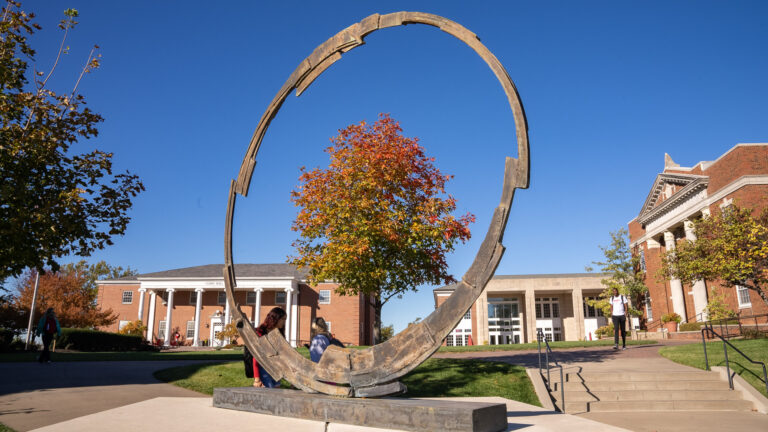 Large bronze ring sculpture on the William Jewell College campus framed by autumn trees with Curry Hall and Yates Gill College Center in the background.