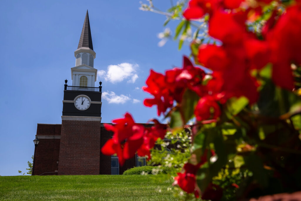 Gano Chapel clock tower at William Jewell College framed by vibrant red flowers on a bright summer day.