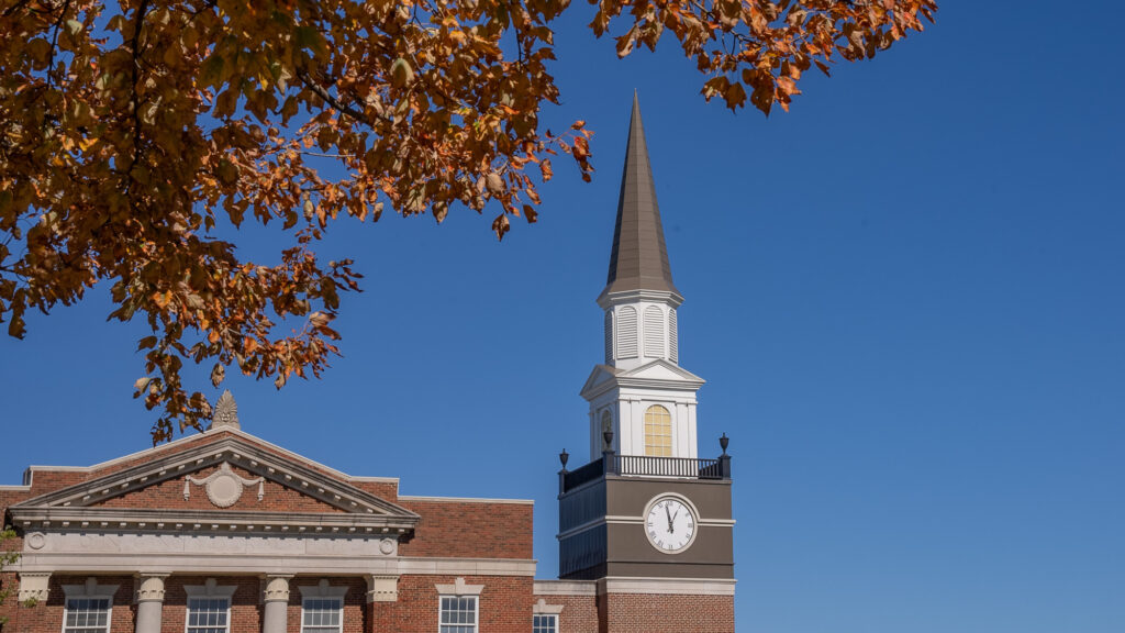 Gano Chapel clock tower spire framing autumn foliage against a clear blue sky at William Jewell College.