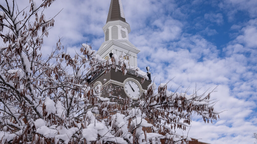 Low-angle view of the snow-dusted Gano Chapel clock tower spire framed by snow-covered tree branches.