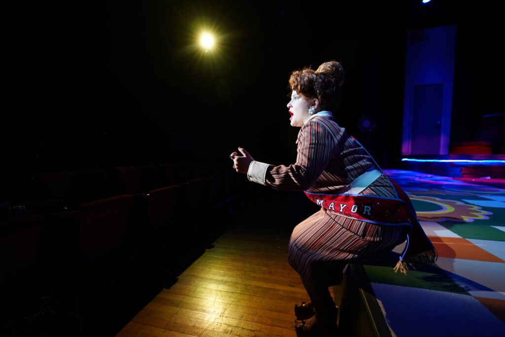 A student actor wearing a "MAYOR" sash gesturing offstage while performing in "The Inspector General."