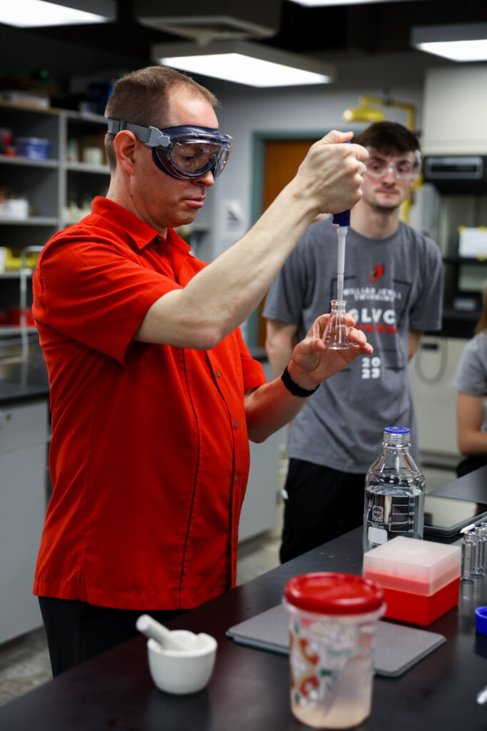 Dr. Shane Price wears safety goggles and a red button-down shirt, demonstrating pipette technique into a glass flask while a student watches in the background.