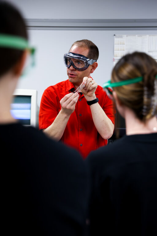 Dr. Shane Price, wearing protective safety goggles, holds a small glass tube while speaking to students in a laboratory.
