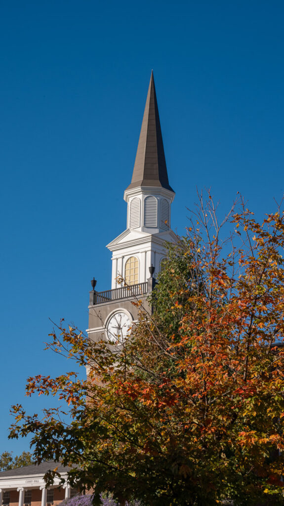 The clock tower spire of Gano Chapel framed by autumn foliage against a blue sky.