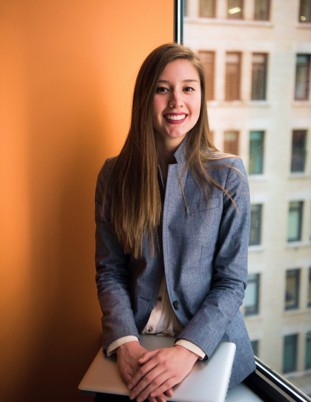 A smiling young woman in a grey blazer resting her hands on a closed laptop near a window overlooking city buildings.