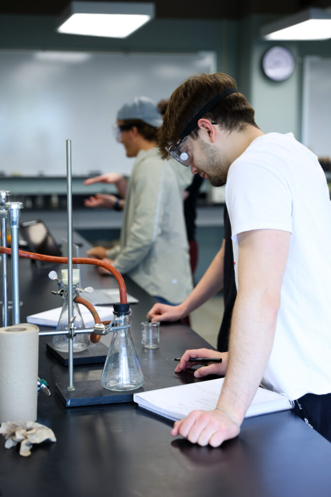 A male student in a white t-shirt and safety glasses leaning over a lab table to write in a notebook during a chemistry experiment.