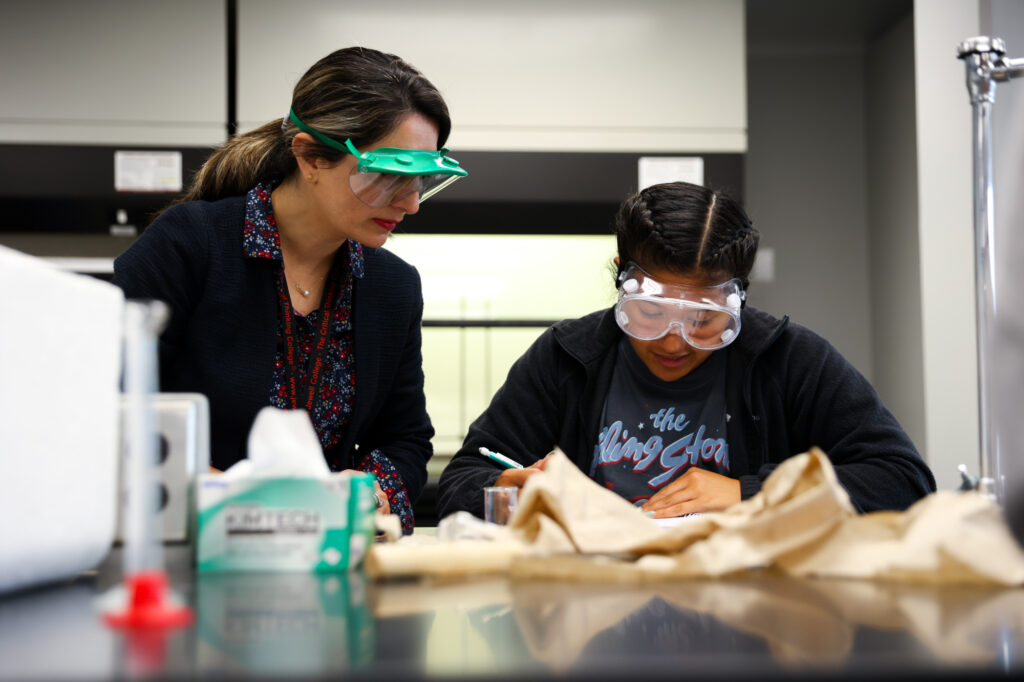 An instructor wearing safety goggles observing a female student in a Rolling Stones t-shirt taking notes at a laboratory work station.
