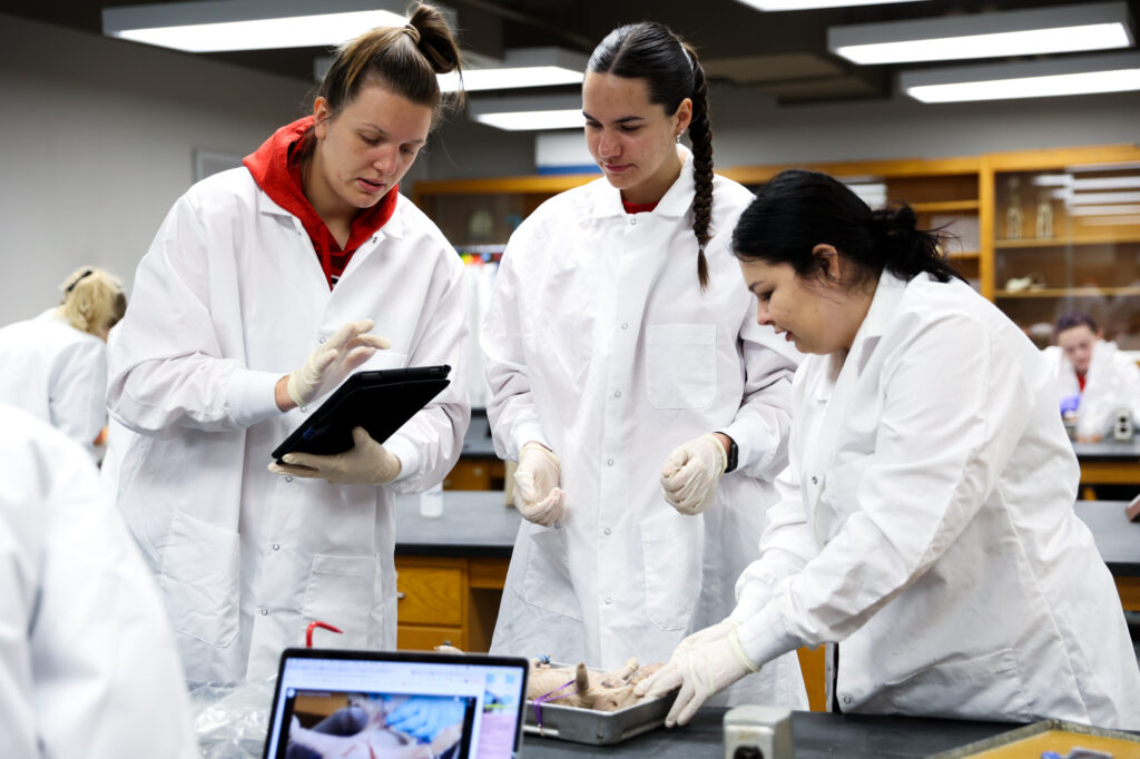 Three female students in white lab coats and gloves reviewing notes on a tablet screen beside a fetal pig dissection specimen in a lab.