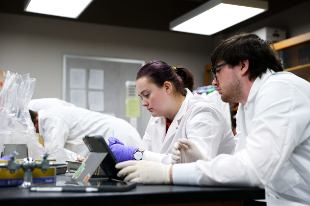 Two students in white lab coats looking down at a tablet screen on a lab bench.