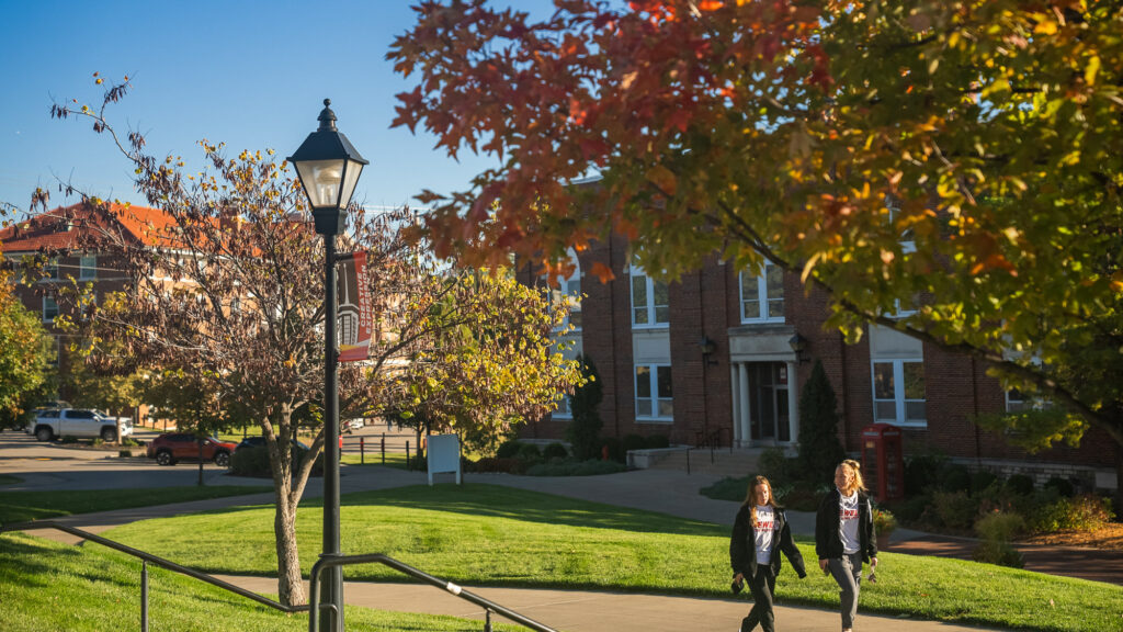 2 students walking along a paved campus pathway past brick buildings and autumn trees.