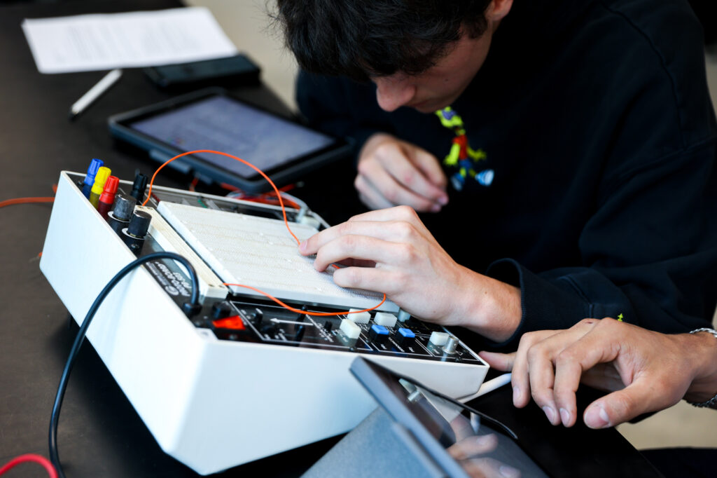 Close-up of a student in a black sweatshirt leaning over a breadboard workstation while connecting an orange circuit wire.