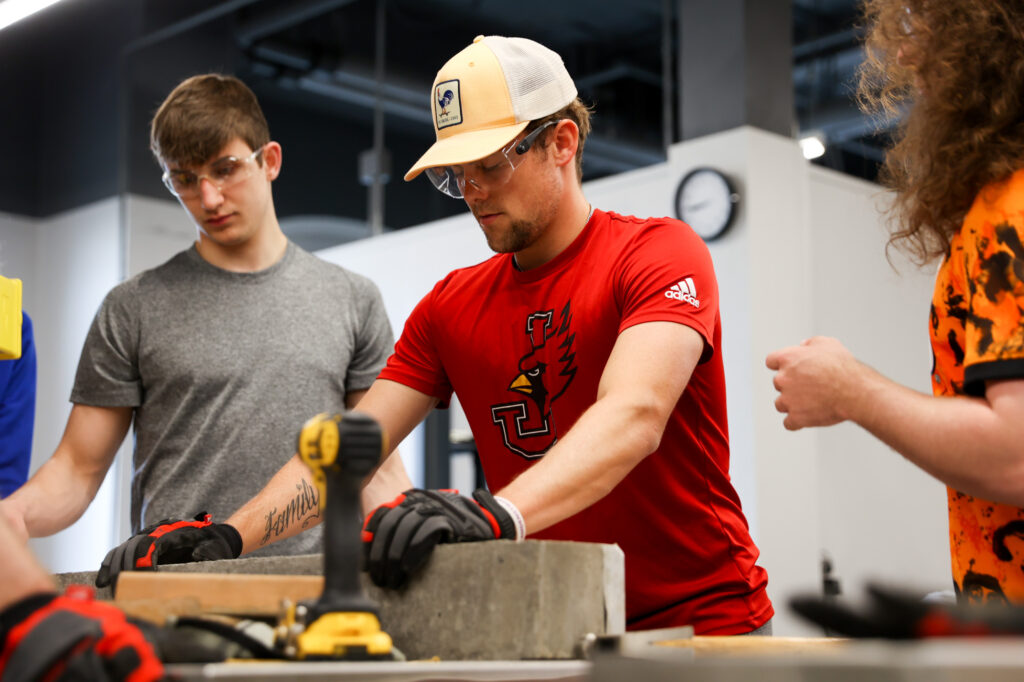 Students wearing safety glasses in a workshop gathered around a lab table holding a concrete beam.