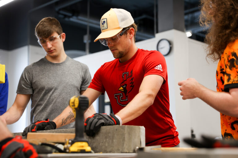 Students wearing safety glasses in a workshop gathered around a lab table holding a concrete beam.