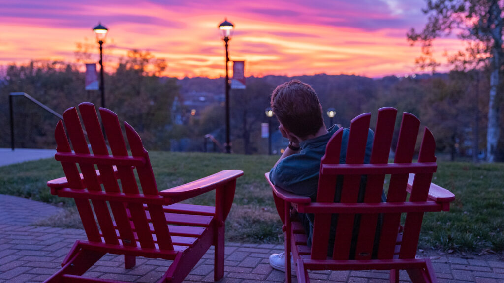 Rear view of 1 person sitting in a red Adirondack chair on campus while watching a vibrant pink and orange sunset.