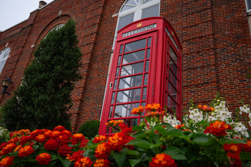 A red British telephone booth standing beside a brick building on campus, framed by colorful garden flowers.