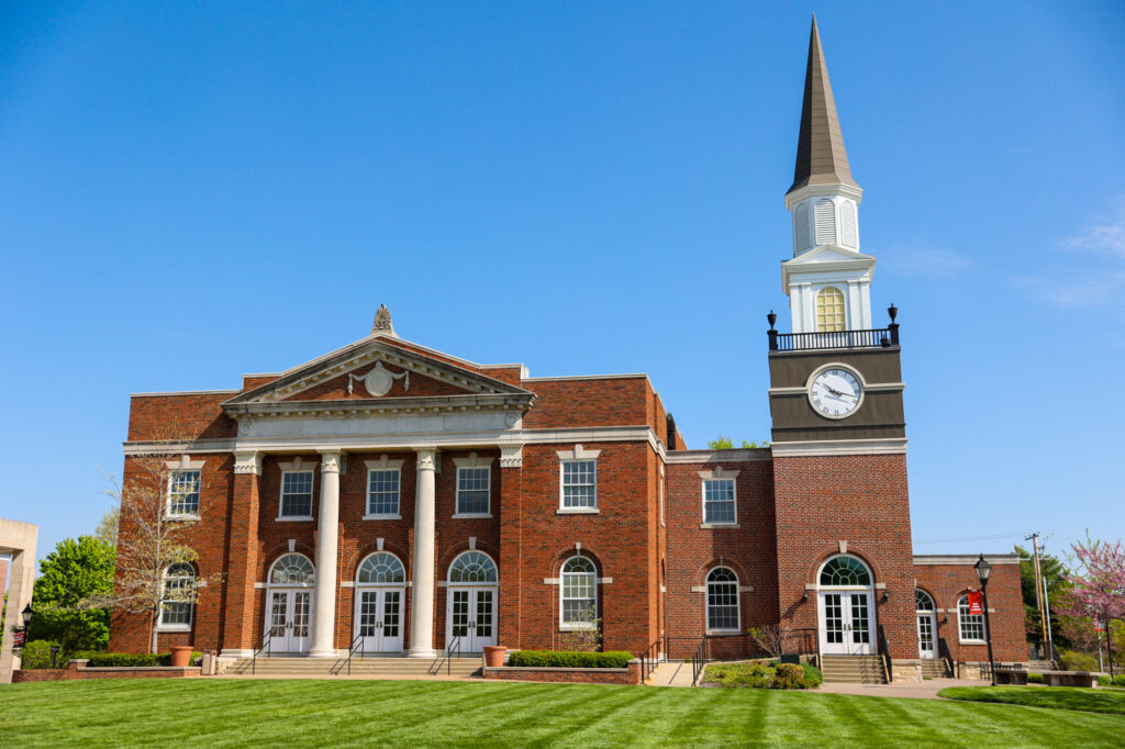 Exterior view of Gano Chapel at William Jewell College, featuring a red brick facade with white columns and a prominent clock tower spire against a clear blue sky.