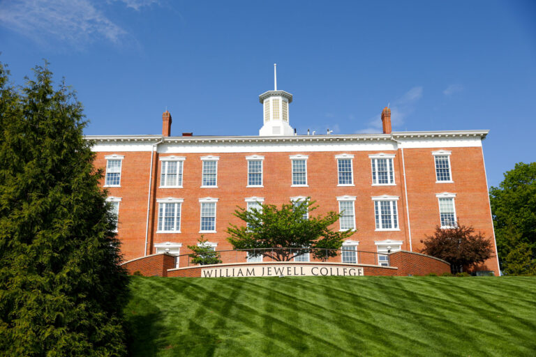 Exterior front view of Jewell Hall at William Jewell College, featuring a red brick facade, cupola, and sloping green lawn displaying the college name.