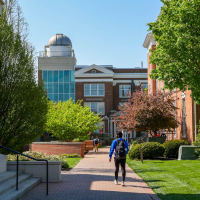 A student wearing a backpack walks along a paved brick campus pathway toward campus buildings, featuring a dome observatory on the left under a clear blue sky.
