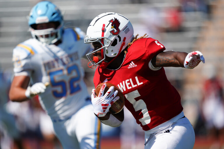 A William Jewell College football player carrying the ball downfield while evading a Fort Lewis College defender.