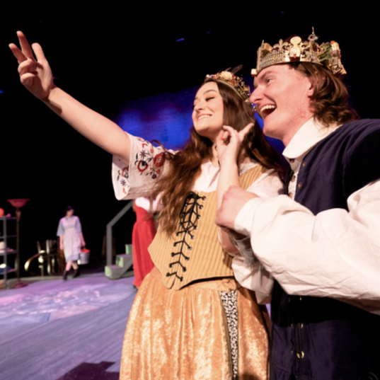 Two student actors wearing crowns and period costumes smiling while taking a picture onstage during a theatrical performance.
