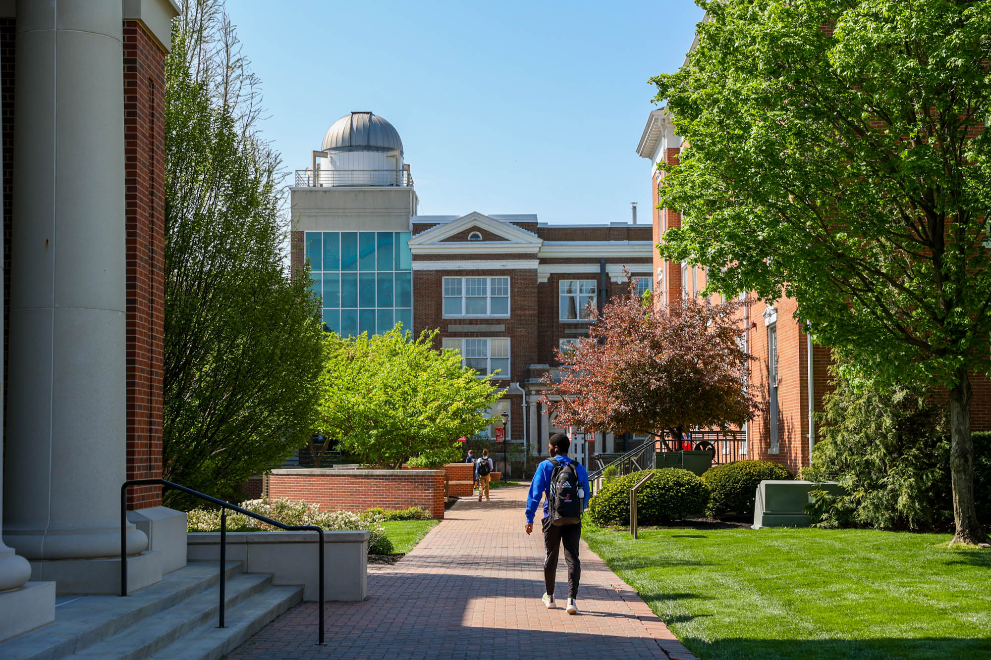 A student wearing a backpack walks along a paved brick campus pathway toward campus buildings, featuring a dome observatory on the left under a clear blue sky.