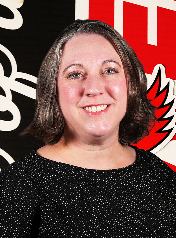 A woman with shoulder-length dark hair and gray highlights smiling in a black polka-dot top, posing for a portrait against a William Jewell athletic backdrop.