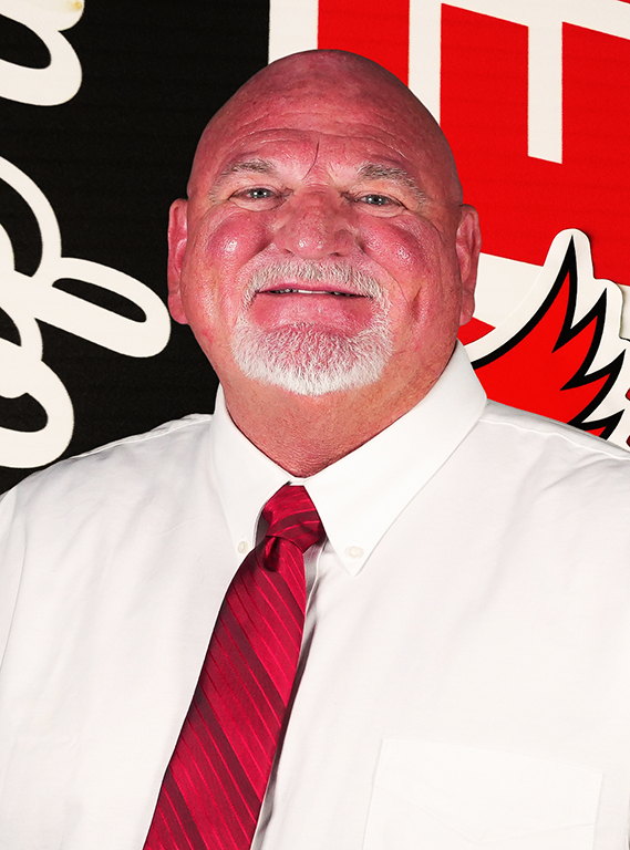 A bald man with a white goatee and mustache smiling in a white dress shirt and red striped tie, posing for a portrait against a William Jewell athletic backdrop.
