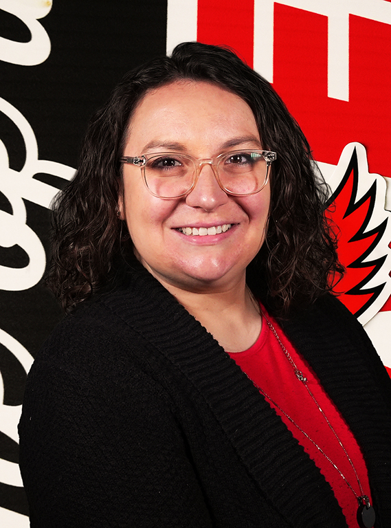 A woman with dark shoulder-length wavy hair wearing clear-framed glasses, a red shirt, and a black cardigan, smiling in front of a William Jewell backdrop.