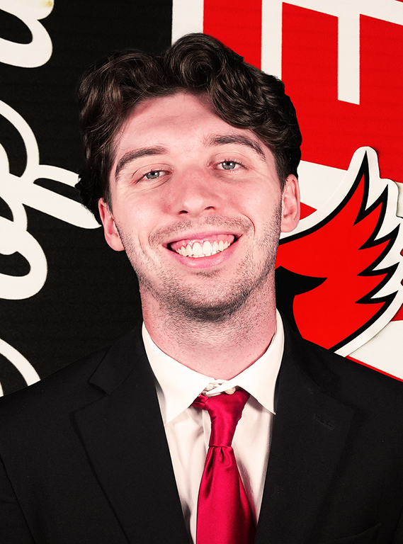 A young man with dark wavy hair smiling in a black suit, white dress shirt, and bright red tie, posing in front of a William Jewell backdrop.