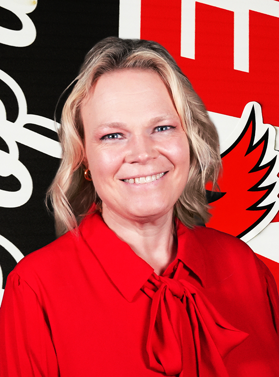 A blonde woman with short wavy hair smiling in a bright red tie-neck blouse, posing in front of a William Jewell backdrop.