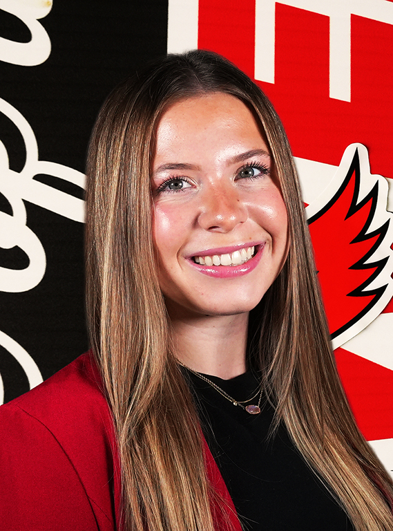 A young woman with long, straight light-brown hair smiling in a black top and red blazer, posing in front of a William Jewell athletic backdrop.
