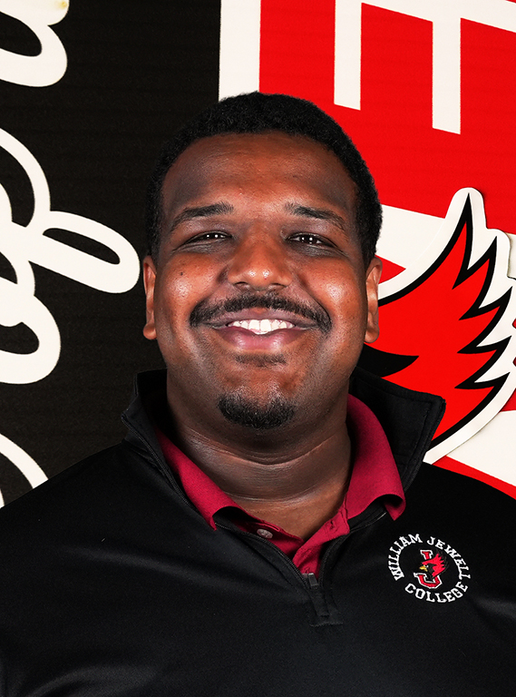 A man with short dark hair, a mustache, and a goatee smiling in a black William Jewell quarter-zip pullover over a red collared shirt, posing in front of an athletic backdrop.