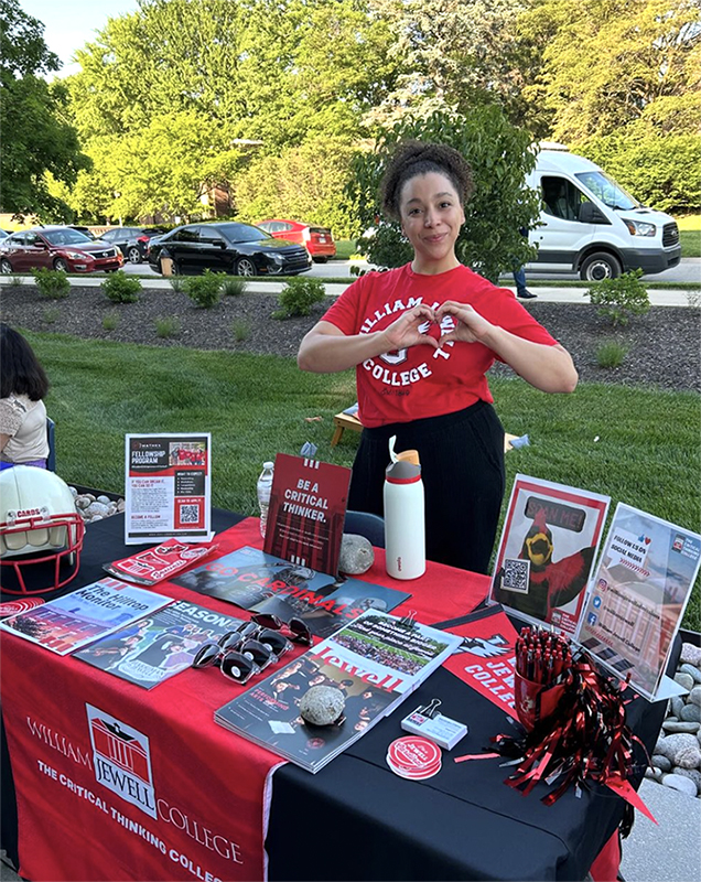 Alexis Morris, Admission Counselor at William Jewell College, making a heart hand gesture while standing behind an admissions table outdoors.