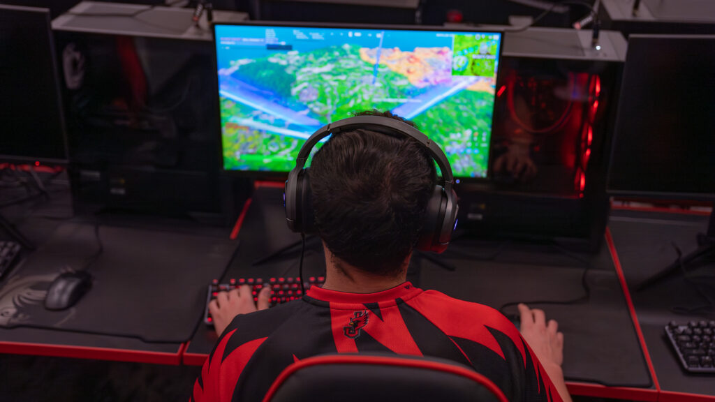 Rear view of an esports student wearing headphones while playing a computer game on a monitor setup in an arena.
