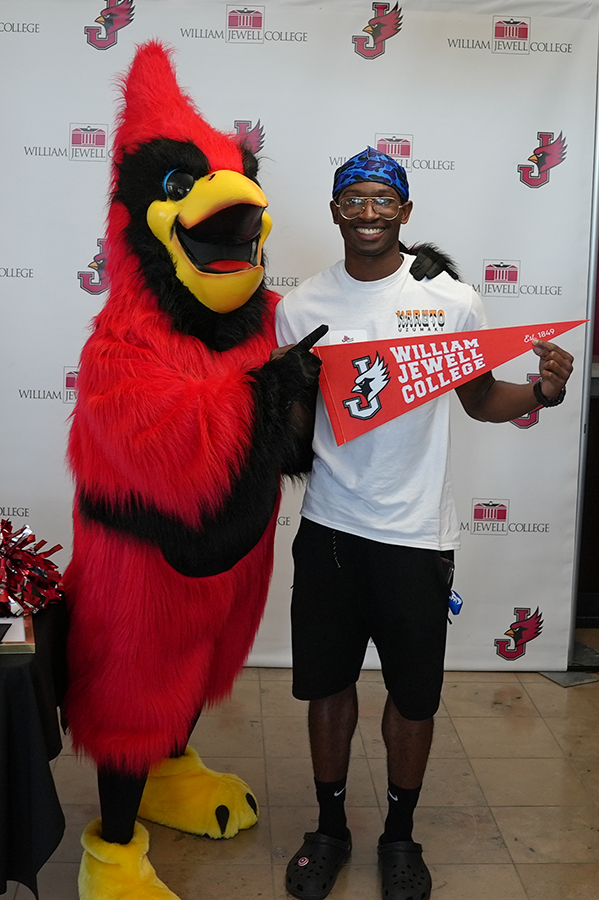 A smiling student posing alongside the William Jewell College Cardinal mascot while holding a college felt pennant.
