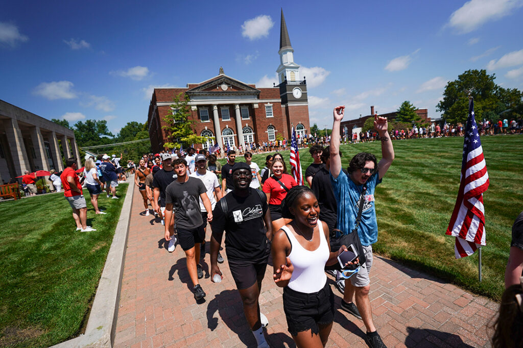 A group of incoming students smiling and walking along a brick pathway across the campus quad in front of Gano Chapel.