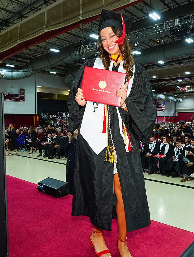 A smiling graduate in a cap, gown, and honors stole holding her red William Jewell College diploma cover inside Mabee Center.