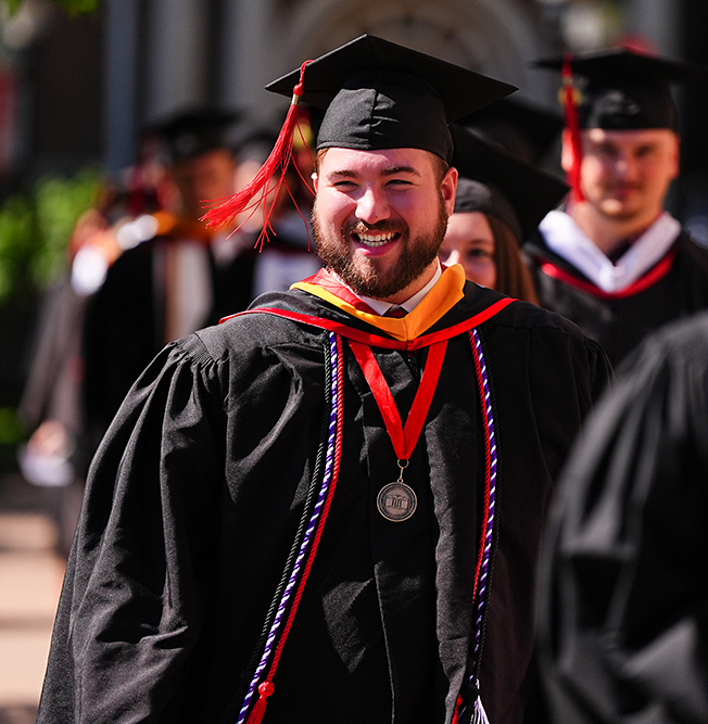 Cameron Huett smiling while walking in the commencement procession in full academic regalia and a medallion.