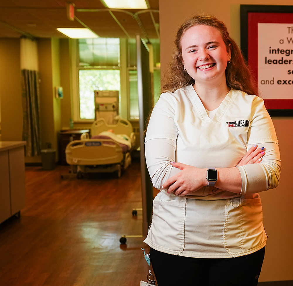 A smiling nursing student in a white William Jewell Nursing scrub top posing with arms crossed in a clinical simulation lab.