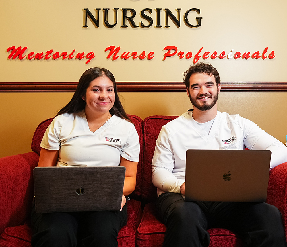 2 nursing students in white William Jewell Nursing scrub tops sitting on a red couch with laptops beneath a wall sign reading "Mentoring Nurse Professionals."