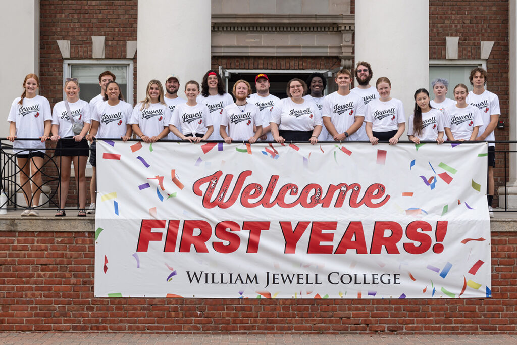Orientation leaders in matching white shirts standing behind a "Welcome FIRST YEARS!" banner on the steps of a campus building.