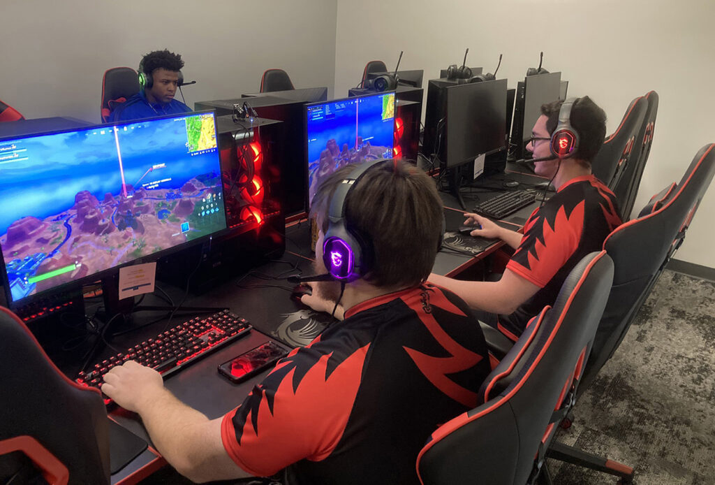 Students in red-and-black jerseys sit at individual computer stations, playing games in an esports lab.
