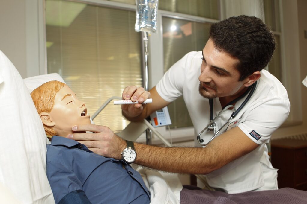 A male nursing student using a penlight to examine the throat of a pediatric mannequin in a hospital bed.